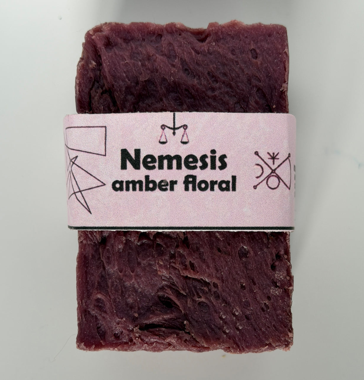 Deep burgundy-maroon handmade soap bar with a porous, textured surface, wrapped in a pale lavender band printed with "Nemesis – amber floral" and geometric mystical symbols
