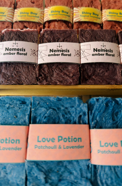 Bouncing Soaps handmade bar soaps displayed on shelf including pink Shiny Boy Tropical Gourmand top row purple Nemesis Amber Floral middle row and blue Love Potion Patchouli and Lavender bottom row