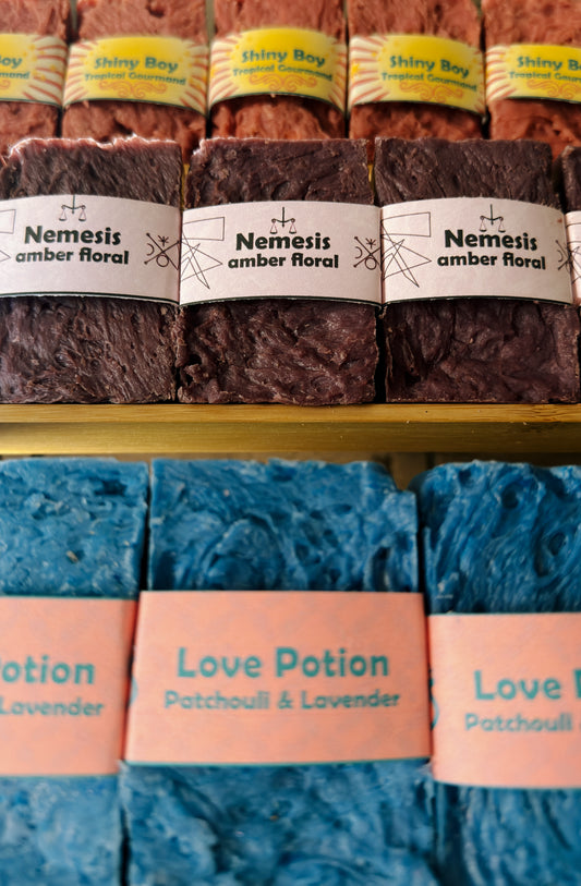 Bouncing Soaps handmade bar soaps displayed on shelf including pink Shiny Boy Tropical Gourmand top row purple Nemesis Amber Floral middle row and blue Love Potion Patchouli and Lavender bottom row