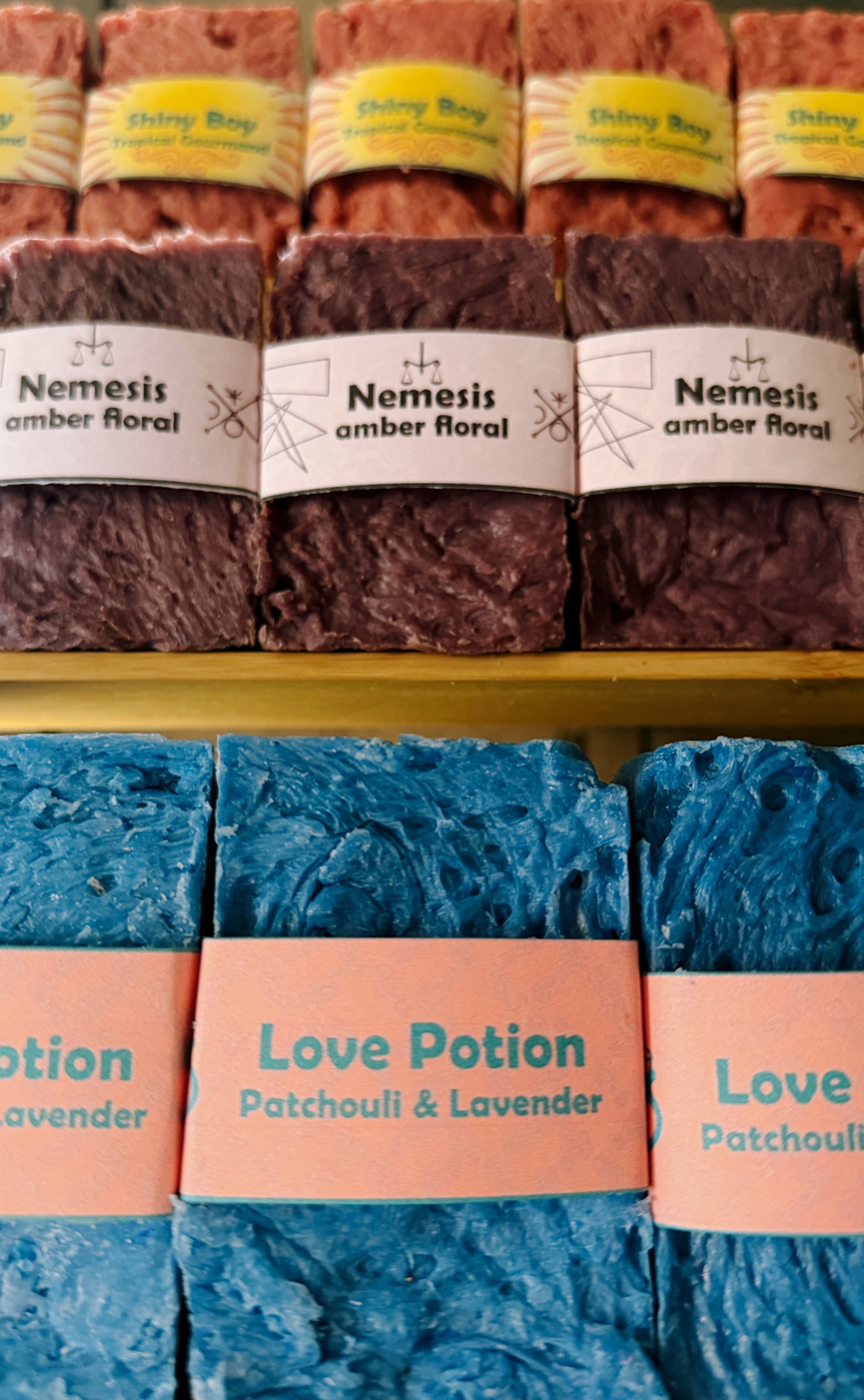 Bouncing Soaps handmade bar soaps displayed on shelf including pink Shiny Boy Tropical Gourmand top row purple Nemesis Amber Floral middle row and blue Love Potion Patchouli and Lavender bottom row