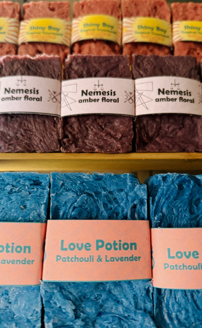 Bouncing Soaps handmade bar soaps displayed on shelf including pink Shiny Boy Tropical Gourmand top row purple Nemesis Amber Floral middle row and blue Love Potion Patchouli and Lavender bottom row