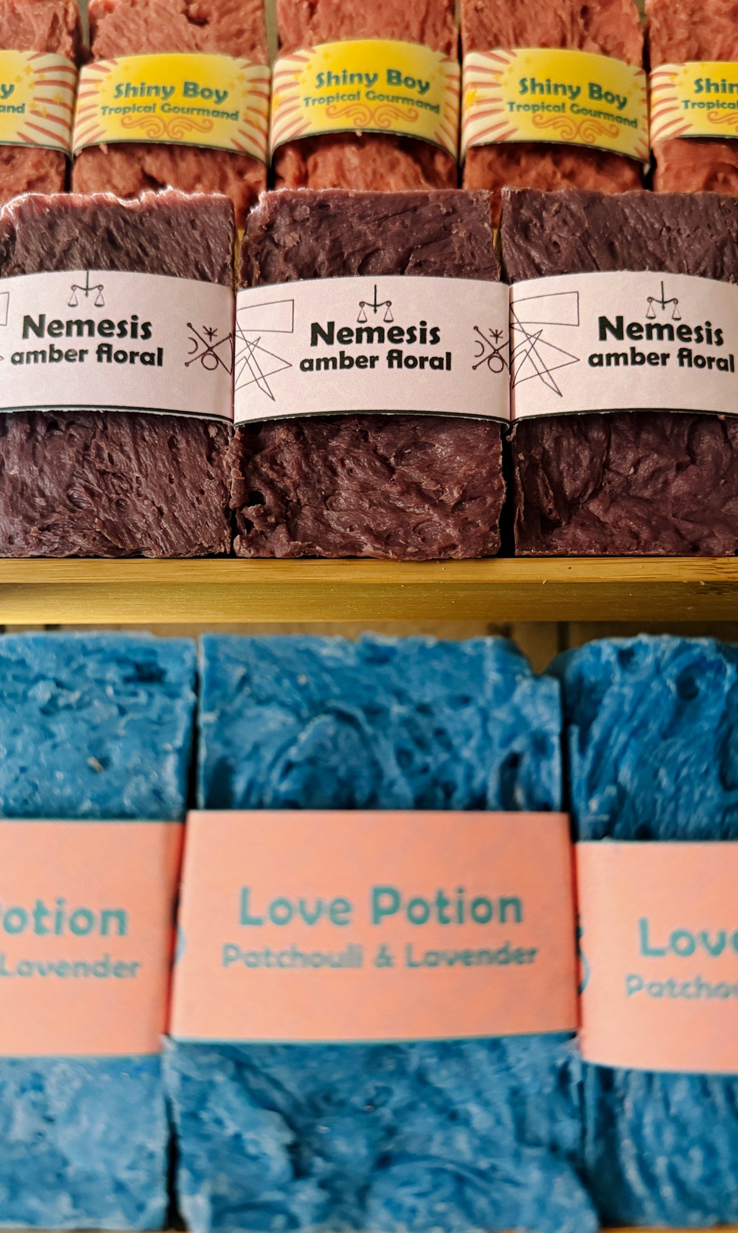 Bouncing Soaps handmade bar soaps displayed on shelf including pink Shiny Boy Tropical Gourmand top row purple Nemesis Amber Floral middle row and blue Love Potion Patchouli and Lavender bottom row