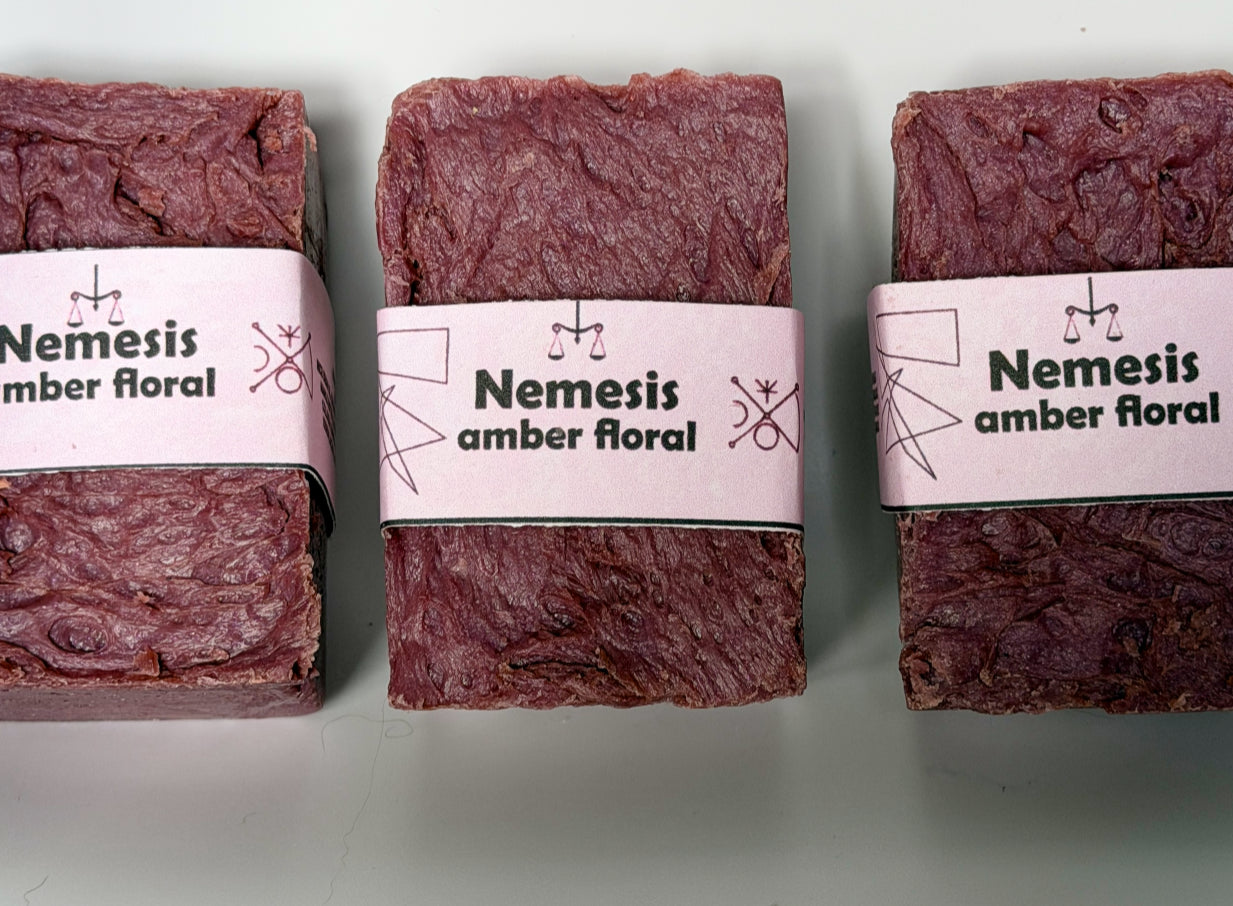 Three Nemesis soap bars arranged in a triangular grouping on a white background, showing the rich maroon color and lavender label from multiple angles
