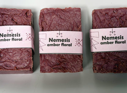 Three Nemesis soap bars arranged in a triangular grouping on a white background, showing the rich maroon color and lavender label from multiple angles