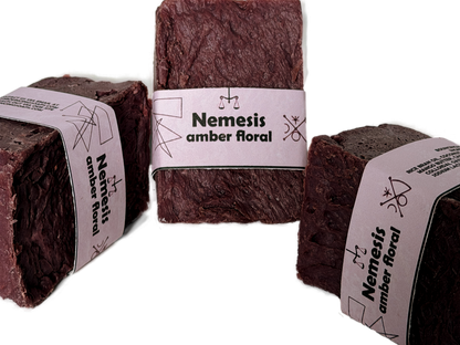 Three Nemesis soap bars arranged in a triangular grouping on a white background, showing the rich maroon color and lavender label from multiple angles