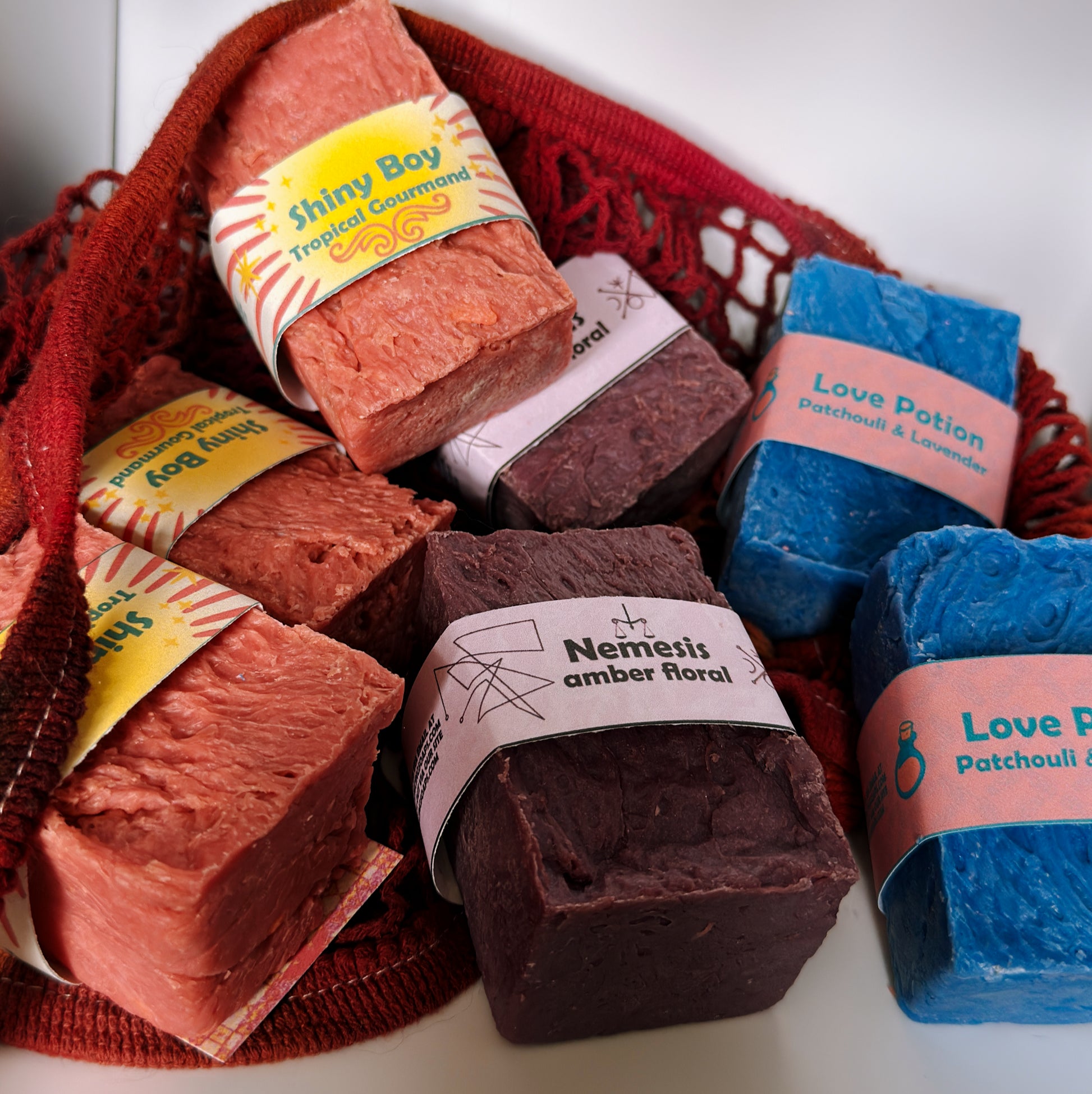 Assorted Bouncing Soaps bars displayed in a crimson knit mesh bag: Shiny Boy (coral), Nemesis (maroon), and Love Potion (blue) bars visible with their branded labels