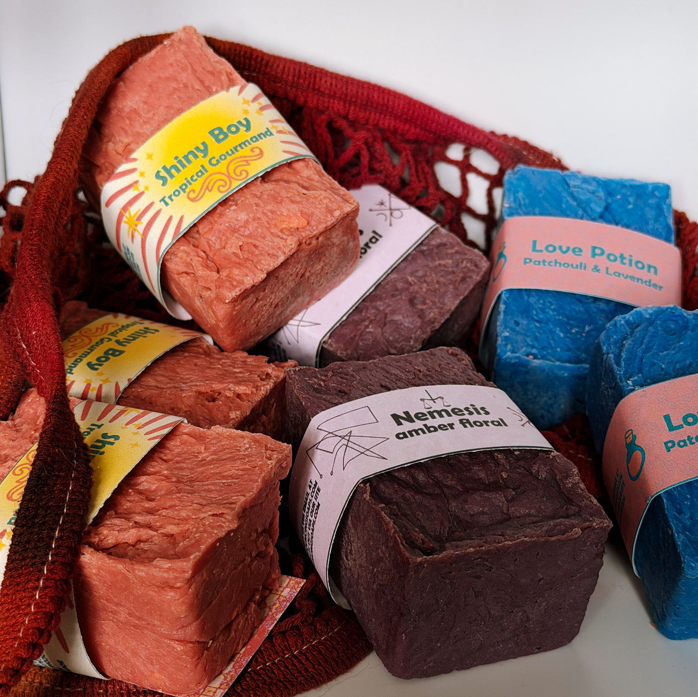 Assorted Bouncing Soaps bars displayed in a crimson knit mesh bag: Shiny Boy (coral), Nemesis (maroon), and Love Potion (blue) bars visible with their branded labels