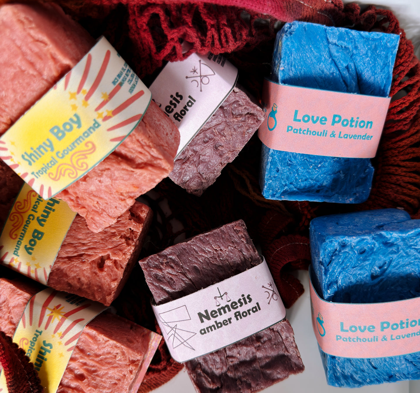 Mixed variety — close-up in red mesh bag
Close-up of Nemesis, Love Potion, and Shiny Boy soap bars nestled in a red mesh bag, highlighting the handcrafted textures and colorful label designs