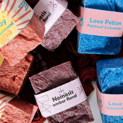 Close-up of Nemesis, Love Potion, and Shiny Boy soap bars nestled in a red mesh bag, highlighting the handcrafted textures and colorful label designs