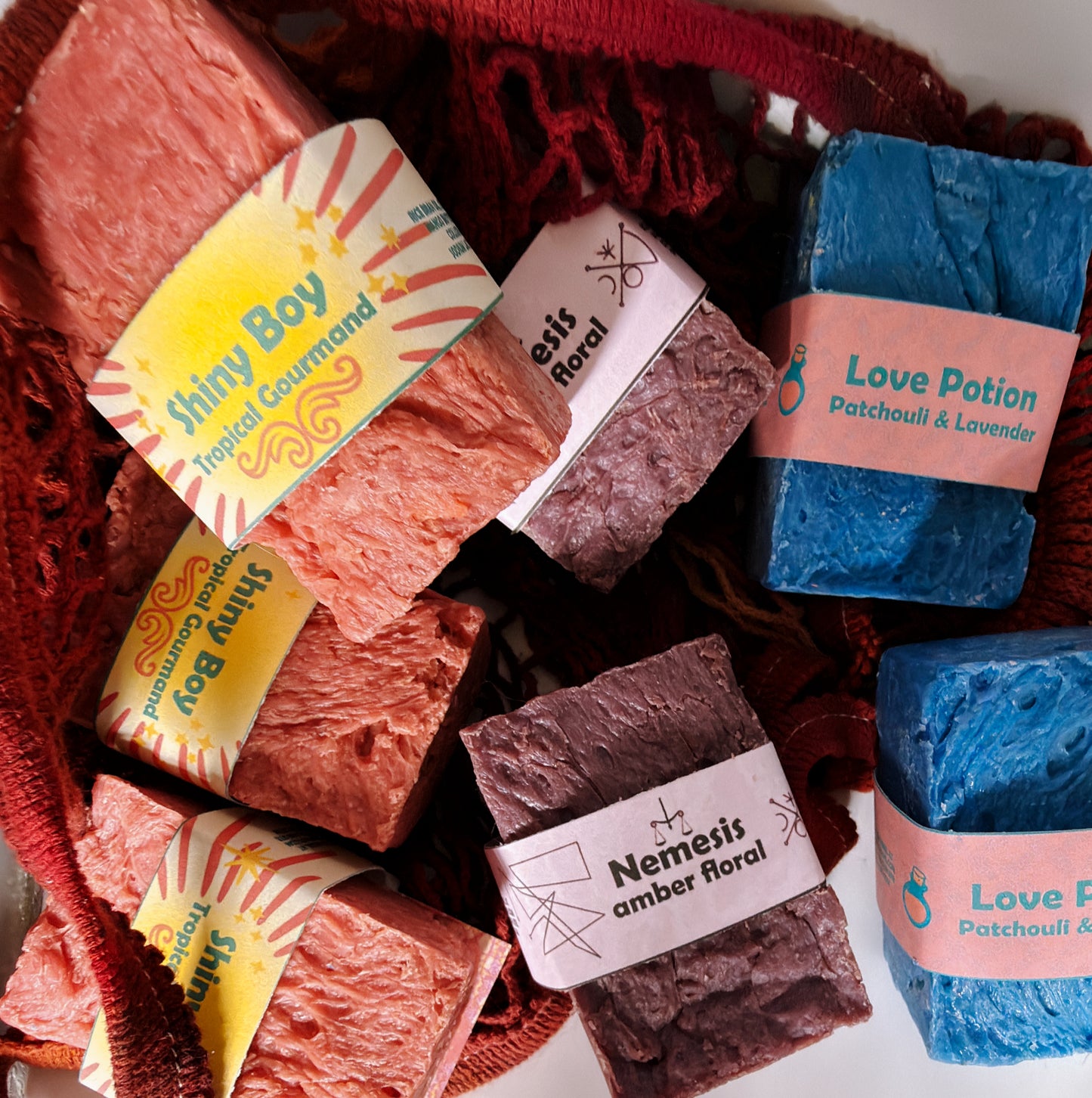 Assorted Bouncing Soaps bars displayed in a crimson knit mesh bag: Shiny Boy (coral), Nemesis (maroon), and Love Potion (blue) bars visible with their branded labels