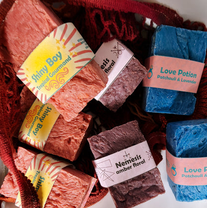 Assorted Bouncing Soaps bars displayed in a crimson knit mesh bag: Shiny Boy (coral), Nemesis (maroon), and Love Potion (blue) bars visible with their branded labels