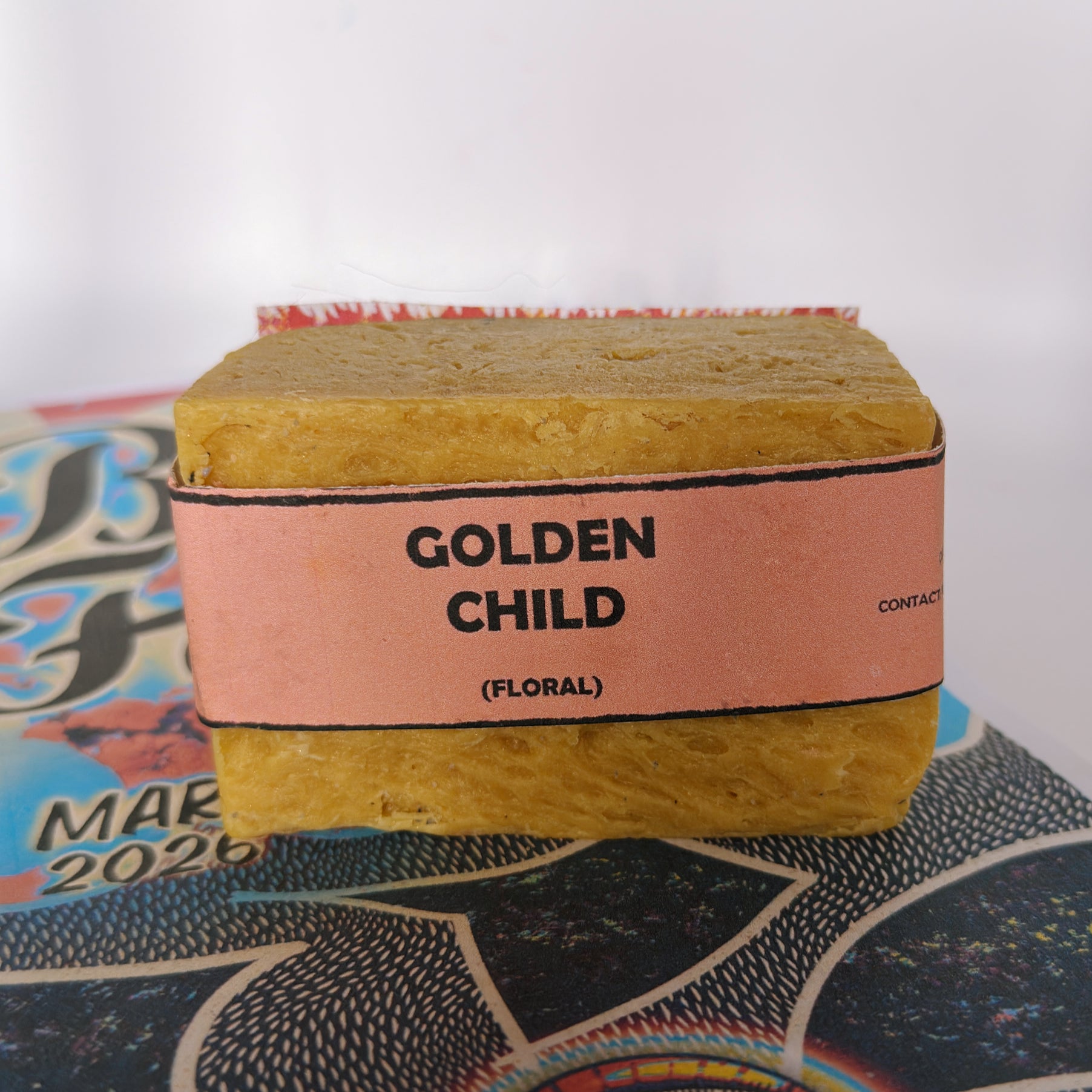 Single bar of Golden Child floral handmade soap by Bouncing Soaps with golden yellow color and pink label resting on Bouncing Soaps branded booklet