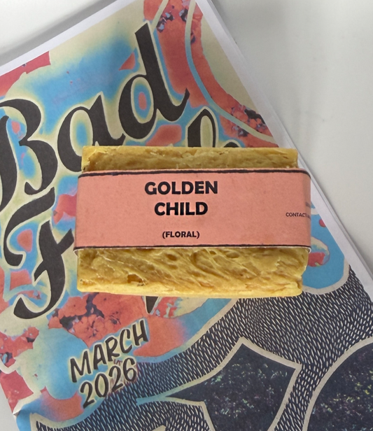 Single bar of Golden Child floral handmade soap by Bouncing Soaps with golden yellow color and pink label resting on Bouncing Soaps branded booklet